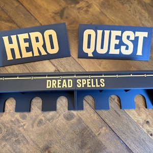 May include: Two dark blue rectangular signs with the text HERO and QUEST in gold lettering. Below them, two dark blue game board organizers with gold text reading DREAD SPELLS and a numbered scale from one to six, featuring arched cutouts.