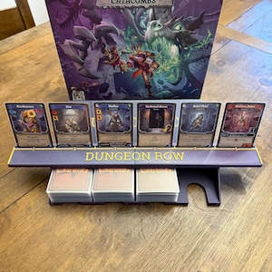 May include: A board game setup featuring a purple "Dungeon Row" card holder with six character cards. The game box, titled "Catacombs," is visible in the background. Several stacks of cards are below the holder, ready for play.