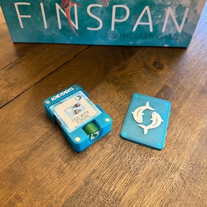 Könnte beinhalten: Eine türkisfarbene Brettspielkomponente mit dem Text "ACHIEVEMENTS" und "FISH WITH TOKENS". Eine passende türkisfarbene Karte mit zwei weißen Fischen befindet sich neben der Komponente. Die Spielbox mit der Aufschrift "FINSPAN A WINGSPAN GAME" befindet sich im Hintergrund.