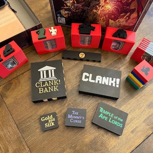 May include: A board game set with various components, including red card holders, black and white game pieces, and game cards. The game pieces include a number 7, and the game titles "CLANK! BANK", "CLANK!", "Gold and Silk", "The Mummy's Curse", and "Temple of the Ape Lords".