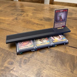 May include: A black plastic game card organizer featuring a horizontal slot for a single upright card labeled GOBLIN and three lower compartments holding stacks of playing cards. The base includes small numbered markers and colored icons.