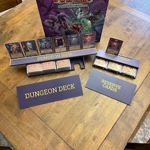 May include: A board game setup with cards and accessories. The game box is visible in the background. The cards are displayed in holders labeled "Dungeon Row" and "Underworld Guardian". Additional cards are in boxes labeled "Dungeon Deck" and "Reserve Cards".