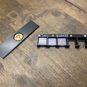 May include: Black game board organizer with a gold coin and the number 7. The organizer has slots for game pieces and the word "MARKET". The game pieces have images of a crown, coins, and a treasure chest.