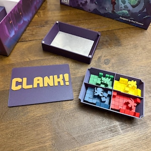May include: A purple board game box with the word "CLANK!" printed on it. The box is open and shows a purple tray with four compartments. Each compartment contains a different colour of wooden cubes and a small plastic figure.