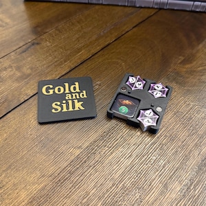 May include: A black box with the words "Gold and Silk" printed in gold letters. The box is open and reveals a small, black tray with four dice and a card with the number 5 on it.