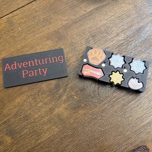 May include: A black rectangular sign with the words "Adventuring Party" in red lettering. Next to it is a black tray with various game pieces, including a paw print, sword, and other geometric shapes in different colors.