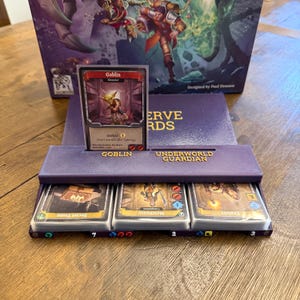 May include: A purple card holder with the text "UNDERWORLD GUARDIAN" and "GOBLIN" holds game cards. The cards feature illustrations and text. A game box with artwork is in the background.