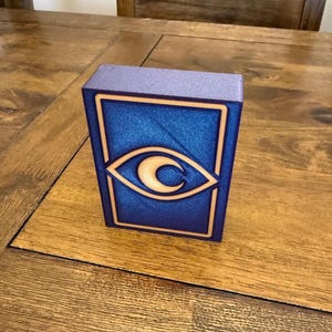 May include: A rectangular box with a blue front featuring a gold-colored eye design. The box has a textured surface and a purple top. The eye design is a stylized crescent moon within an eye shape.