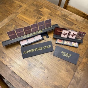 May include: A black plastic card organizer set for a tabletop game, featuring two long card stands with gold text reading ADVENTURE ROW and CULTIST. Two rectangular black signs with gold text read ADVENTURE DECK and RESERVE CARDS, with stacks of game cards.