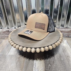 May include: A tan and black trucker hat with a mesh back. The front of the hat features a leather patch with the text "MOUNTAIN LION" and "COUGAR". The hat is displayed on a wooden beaded tray.