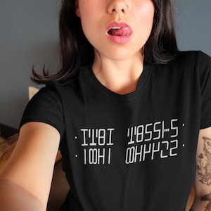 Może przedstawiać: Czarny t-shirt z krótkim rękawem z białym nadrukiem na klatce piersiowej. Tekst jest ułożony w dwóch liniach i wygląda na lustrzane odbicie, czytając "IT'S OKAY I'M LOST" w stylizowanej, blokowej czcionce. Koszulka ma okrągły dekolt.