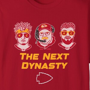 May include: Red t-shirt with a graphic of three men's faces. The men are wearing sunglasses and have the letters "K.C." on their sunglasses. The text "THE NEXT DYNASTY" is below the faces. A red arrowhead is below the text.