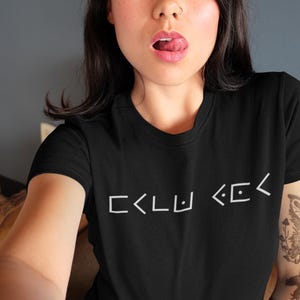 Puede incluir: Una camiseta negra con un gráfico blanco del texto "CKLU KCK" en la parte delantera.