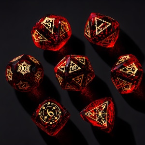 Puede incluir: Un juego de siete dados poliédricos de color rojo translúcido con bordes afilados y grabados dorados intrincados. Cada dado presenta patrones geométricos y números únicos, incluyendo un dado de veinte caras y uno de doce, dispuestos sobre una superficie oscura.
