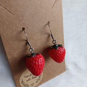 Op de afbeelding: Een paar rode aardbei oorhangers met zilveren haakjes. De oorhangers zijn op een bruine kaart met de tekst "handgemaakt met liefde" en een klein hartje.