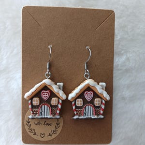 Könnte beinhalten: Ein Paar Ohrringe in Form eines Lebkuchenhauses mit weißem Zuckerguss, roten Zuckerstangen und einer braunen Tür. Die Ohrringe befinden sich auf einer braunen Karte mit dem Text "with love" in einem Kreis.