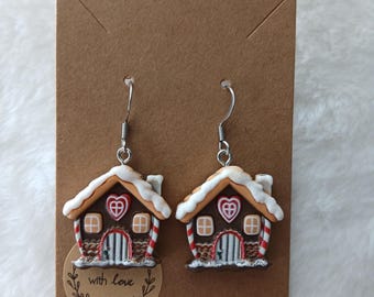 May include: A pair of gingerbread house earrings with white icing, red candy canes, and a brown door. The earrings are on a brown card with the text "with love" in a circle.