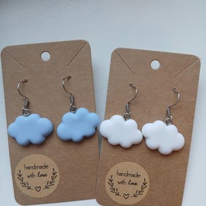 Peut inclure: Deux paires de boucles d'oreilles faites à la main avec un motif de nuage. Une paire est bleu clair et l'autre paire est blanche. Les boucles d'oreilles sont présentées sur du carton brun avec un autocollant rond qui dit "fait main avec amour".