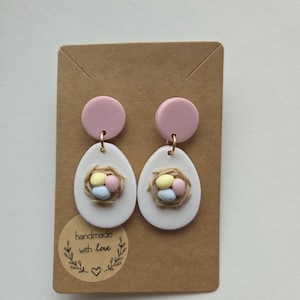 Könnte beinhalten: Ein Paar Ohrringe zum Thema Ostern. Jeder Ohrring hat einen rosa runden Ohrstecker, einen weißen eiförmigen Anhänger mit einem Nest aus pastellfarbenen Eiern. Auf der Karte steht der Text "handmade with love".