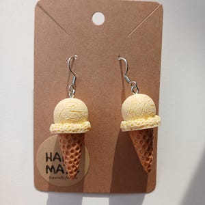 May include: A pair of novelty earrings shaped like ice cream cones. Each earring features a light yellow ice cream scoop atop a brown waffle cone. The earrings hang from silver-colored fish hook ear wires. The earrings are displayed on a brown card.