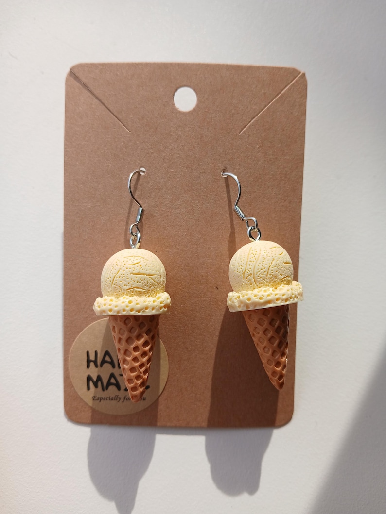 May include: A pair of novelty earrings shaped like ice cream cones. Each earring features a light yellow ice cream scoop atop a brown waffle cone. The earrings hang from silver-colored fish hook ear wires. The earrings are displayed on a brown card.