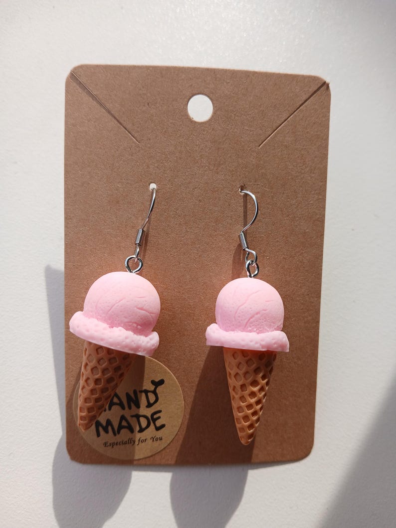 May include: A pair of dangle earrings featuring miniature pink ice cream scoops in waffle cones. The earrings are attached to a brown rectangular display card with a circular sticker that reads HAND MADE Especially for You.