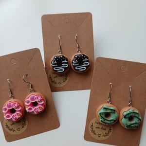Könnte beinhalten: Drei Paare Donut-Ohrringe auf braunem Karton. Die Donuts sind aus Polymer Clay gefertigt und haben verschiedene Beläge. Ein Paar hat einen Schokoladenüberzug mit weißem Drizzle, ein Paar hat rosa Zuckerguss mit bunten Streuseln und ein Paar hat grünen Zuckerguss mit einem schwarzen Strudel und gelben Streuseln. Jede Karte hat einen kleinen Kreis mit dem Text "Handgefertigt mit Liebe" und einem Herzen.