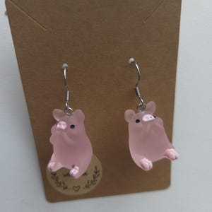 Op de afbeelding: Een paar hangende oorbellen met doorschijnende, matroze varkentjesbedels met kleine zwarte oogjes en roze geschilderde snuiten. De varkentjesfiguren hangen aan zilverkleurige metalen haken en zijn bevestigd aan een rechthoekige bruine kaart.