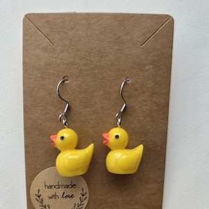 Op de afbeelding: Een paar gele eenden oorbellen met oranje snavels en zilveren haken. De oorbellen worden gepresenteerd op een bruine kaart met een sticker met de tekst "handmade with love".