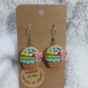 Op de afbeelding: Een paar oorbellen met Pasen als thema. Elke oorbel heeft een miniatuur mandontwerp met kleurrijke eieren, groen gras en een roze strik. De oorbellen zijn bevestigd aan een bruine kaart met de tekst "handmade with love".