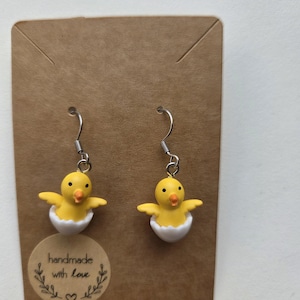 Op de afbeelding: Oorbellen met twee gele kuikens met oranje snavels en uitgespreide vleugels, die uit witte eierschalen komen. De oorbellen hangen aan zilveren haken en worden gepresenteerd op een bruine kaart met de tekst "handmade with love".