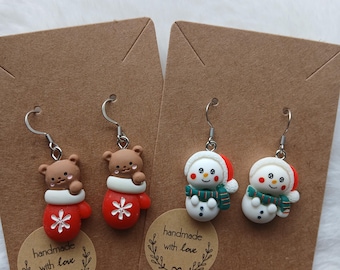 May include: Two pairs of festive earrings. One pair features brown bear figures in red mittens with white snowflake accents. The other pair showcases snowmen with red hats, green scarves, and black button eyes. Each pair is attached to a brown card.