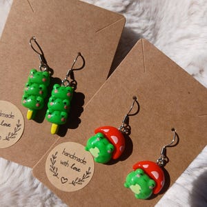 Könnte beinhalten: Handgefertigte Polymer Clay Ohrringe. Ein Paar zeigt grüne, froschförmige Eis am Stiel auf gelben Stäben. Das andere Paar hat grüne Frösche unter rot-weiß gepunkteten Pilzkappen. Beide Paare haben silberfarbene Ohrhaken.