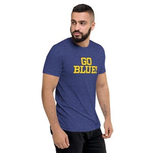 Könnte beinhalten: Ein kurzärmeliges T-Shirt in meliertem Blau mit dem Text GO BLUE!, der in fetten gelben Blockbuchstaben auf der Brust gedruckt ist. Das Kleidungsstück hat einen Rundhalsausschnitt und eine Standardpassform aus weichem, strukturiertem Stoff.