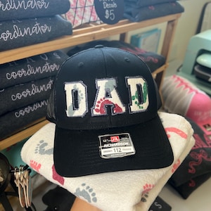 May include: Black trucker hat with the word "DAD" in white block letters with a floral pattern. The hat is sitting on a white towel with gray and pink footprints. The hat is adjustable.