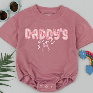 May include: A dusty rose baby romper with short sleeves and a round neckline. The text "DADDY'S girl" is printed on the front in a patchwork design. A small bow is below the text. Light blue sunglasses and a rainbow decoration are also in the image.