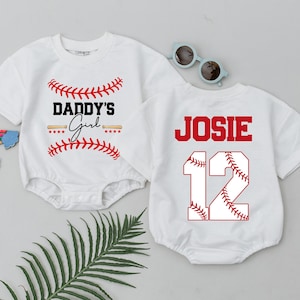 May include: Two white baby onesies with baseball-themed designs. One features "Daddy's Girl" with baseball stitching and stars. The other has "JOSIE" and the number "12" with baseball stitching. Sunglasses and a rainbow decoration are also present.