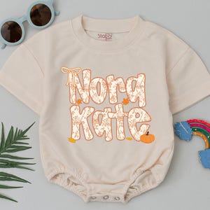 May include: A beige short-sleeved baby romper featuring the text Nora Kate in a floral-patterned font, accented with a small bow graphic and an orange pumpkin. The garment includes a ribbed crew neckline and snap closures at the bottom hem.