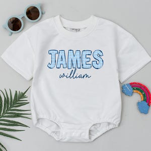 May include: White baby bodysuit with the name "JAMES" in blue gingham print and the name "william" in blue script. The bodysuit has short sleeves and a round neckline. Sunglasses and a rainbow toy are in the background.