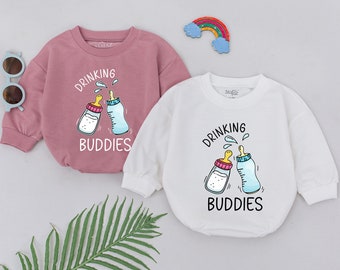 以下が含まれることがあります： 2つのベビーロンパース。2つの哺乳瓶が乾杯しているグラフィックと「Drinking Buddies」というテキストが描かれています。1つのロンパースはピンクで、もう1つは白です。
