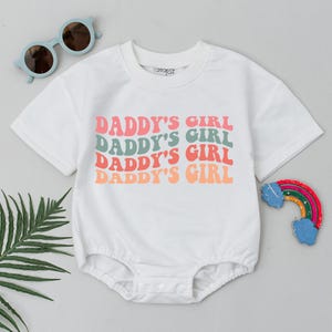 Op de afbeelding: Witte baby romper met de tekst "Daddy's Girl" vier keer herhaald in een retro lettertype met roze, oranje en groene kleuren.