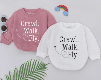 May include: Two baby bodysuits, one dusty rose and one white, with the text "Crawl. Walk. Fly." in black. Each bodysuit has a small airplane graphic with a dotted line leading to a heart. A pair of sunglasses and a rainbow decoration are also visible.