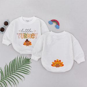 May include: White long-sleeve baby onesie with the text "little TURKEY" in a plaid font and a cartoon turkey illustration. The back of the onesie has a cartoon turkey tail illustration.