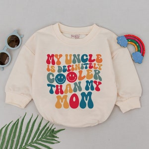 May include: A cream colored sweatshirt with long sleeves and a crew neck. The sweatshirt has a retro-style graphic that says "My Uncle is Definitely Cooler Than My Mom" in colorful letters with smiley faces.