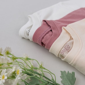 May include: Three folded t-shirts in white, pink, and cream colors. The cream t-shirt has the word "Stofz" printed on it.