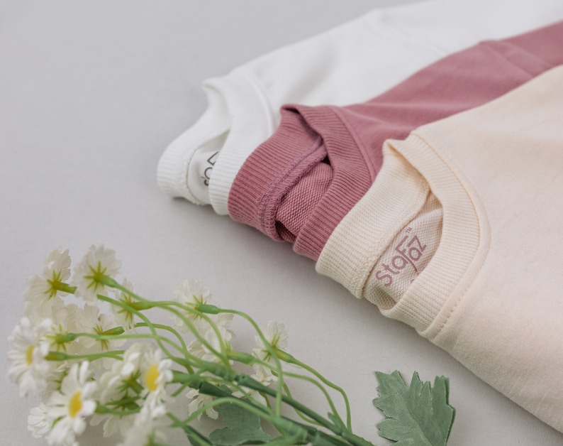 May include: Three folded t-shirts in white, pink, and cream colors. The cream t-shirt has the word "Stofz" printed on it.