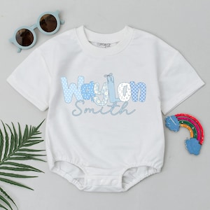 May include: White baby bodysuit with the name "Waylon Smith" in blue lettering. The name is designed with various patterns, including polka dots and checks. Light blue sunglasses and a rainbow-shaped toy are also visible.