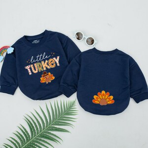 May include: Two navy blue long-sleeved shirts with Thanksgiving-themed designs. One shirt features the text "little TURKEY" with a cartoon turkey in a pilgrim hat. The other shirt has a cartoon turkey tail on the back.