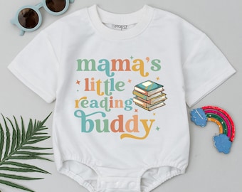 Może przedstawiać: Biały body z krótkim rękawem z napisem "mama's little reading buddy" w kolorowych literach. Obok tekstu znajduje się ilustracja stosu książek.