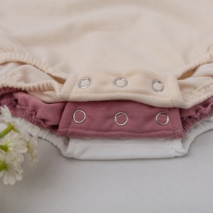 May include: Three baby onesies stacked on top of each other. The top onesie is cream colored, the middle onesie is a dusty rose color, and the bottom onesie is white. All three onesies have silver snaps.